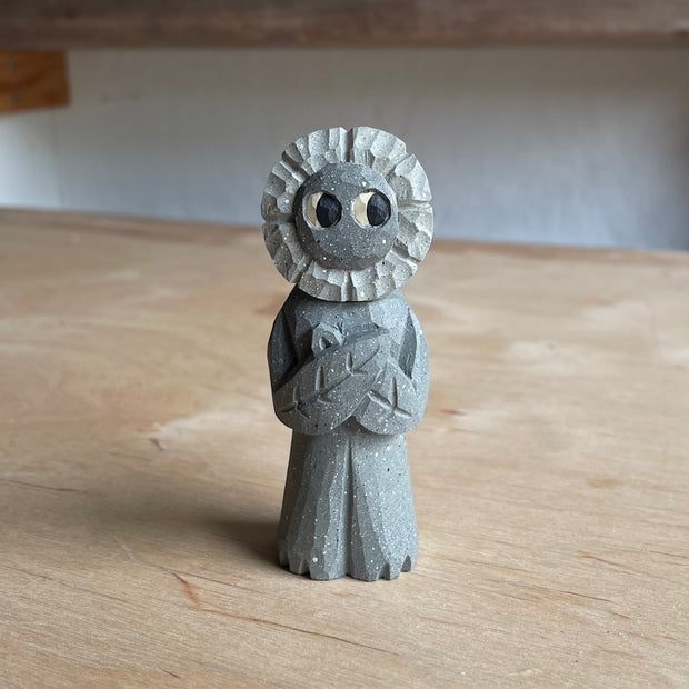 Whittled wooden sculpture of a flower person, with only eyes. It looks off to the side and stands with its leaf arms across its body. It is made to look like it is stone.