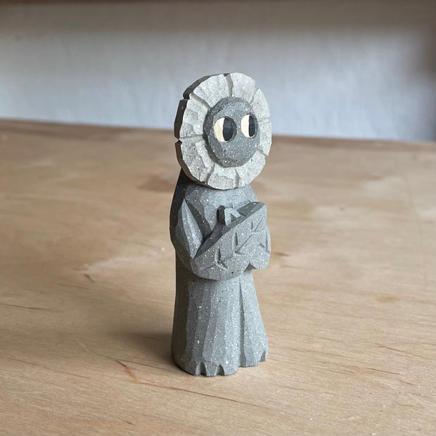 Whittled wooden sculpture of a flower person, with only eyes. It looks off to the side and stands with its leaf arms across its body. It is made to look like it is stone.