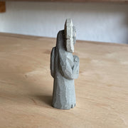 Whittled wooden sculpture of a flower person, with only eyes. It looks off to the side and stands with its leaf arms across its body. It is made to look like it is stone.