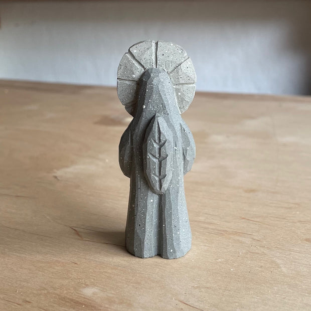 Whittled wooden sculpture of a flower person, with only eyes. It looks off to the side and stands with its leaf arms across its body. It is made to look like it is stone.