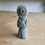 Whittled wooden sculpture of a flower person, with only eyes. It looks off to the side and stands with its leaf arms across its body. It is made to look like it is stone.