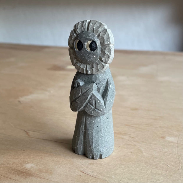 Whittled wooden sculpture of a flower person, with only eyes. It looks off to the side and stands with its leaf arms across its body. It is made to look like it is stone.