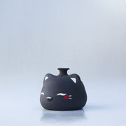 Small, short black jar with a painted on cat face, with closed eyes and red cheeks. It has a small spout opening.