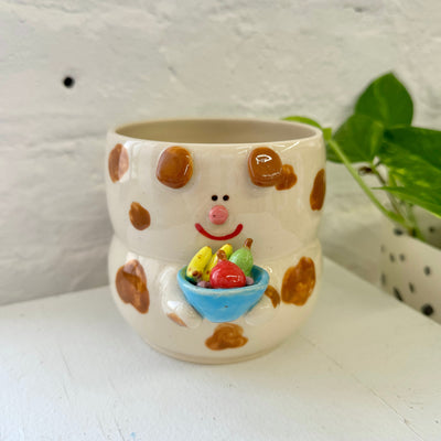 Ceramic cup, light brown made to look like a dog with a cute cartoon smiling face. It holds a blue bowl in its hands that is filled with fruit: bananas, an apple and a pear.