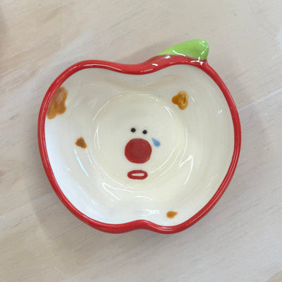 Small ceramic plate, outlined like a red apple in shape and color. Interior of plate is white with small brown splotches and a cartoon face, shedding a small tear. 
