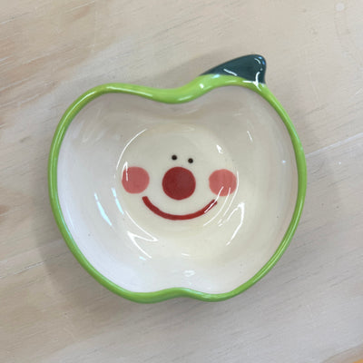 Small ceramic plate, outlined like a green apple in shape and color. Interior of plate is white with a cute, smiling cartoon face.