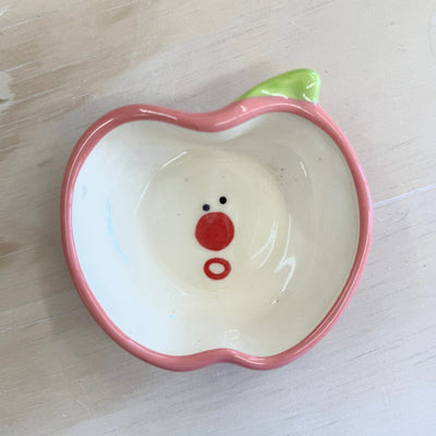 Small ceramic plate, outlined like a pink apple in shape and color. Interior of plate is white with a cute, shocked cartoon face.