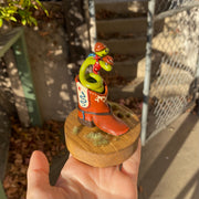 Sculpture of a 2 headed green snake coiled up inside of a brown cowboy boot. Each head of the snake has a brown cowboy hat and a bandana. Boot has decorative elements of a blue snake and red star on each side, with additional cream patterning in the "embroidery"