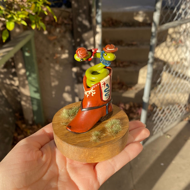 Sculpture of a 2 headed green snake coiled up inside of a brown cowboy boot. Each head of the snake has a brown cowboy hat and a bandana. Boot has decorative elements of a blue snake and red star on each side, with additional cream patterning in the "embroidery"