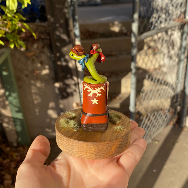 Sculpture of a 2 headed green snake coiled up inside of a brown cowboy boot. Each head of the snake has a brown cowboy hat and a bandana. Boot has decorative elements of a blue snake and red star on each side, with additional cream patterning in the "embroidery"