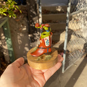 Sculpture of a 2 headed green snake coiled up inside of a brown cowboy boot. Each head of the snake has a brown cowboy hat and a bandana. Boot has decorative elements of a blue snake and red star on each side, with additional cream patterning in the "embroidery".