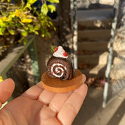 Small sculpture of a snake shaped like a chocolate swiss roll with strawberry cream filling. The snake's head comes out of the cake swirl and has a dollop of whipped cream. Atop the snake's back is a larger dollop of whipped cream with strawberries.