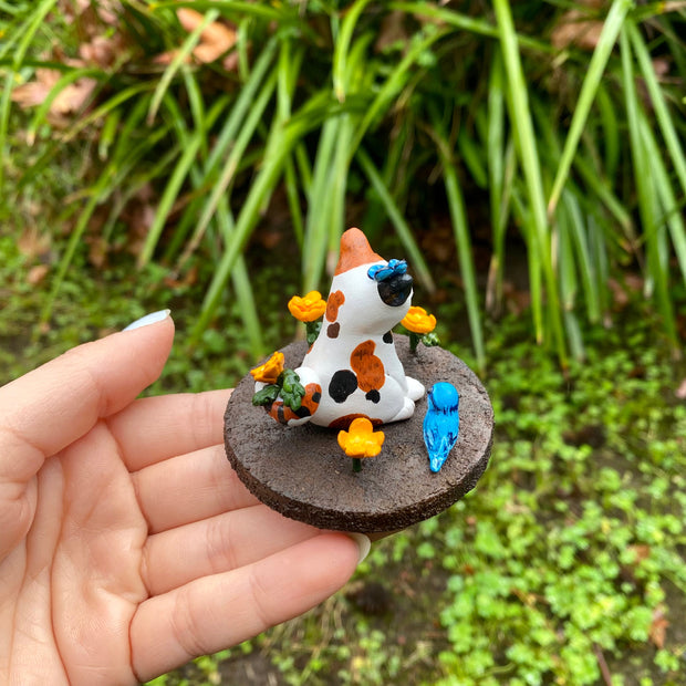 Sculpture of a calico cat sitting with one paw raised and a closed eye, smiling expression. A blue butterfly sits atop its head. The cat holds an orange flower in its tail and sits on a brown dirt circular base with orange flowers and a blue and orange bird.