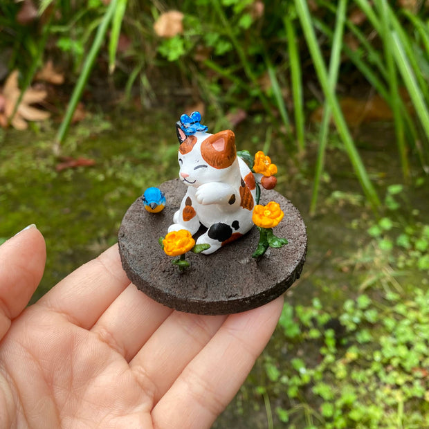 Sculpture of a calico cat sitting with one paw raised and a closed eye, smiling expression. A blue butterfly sits atop its head. The cat holds an orange flower in its tail and sits on a brown dirt circular base with orange flowers and a blue and orange bird.