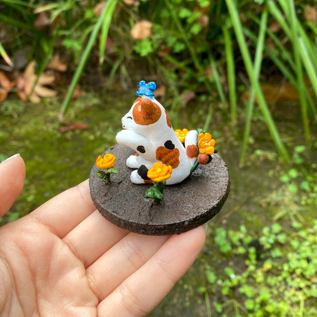 Sculpture of a calico cat sitting with one paw raised and a closed eye, smiling expression. A blue butterfly sits atop its head. The cat holds an orange flower in its tail and sits on a brown dirt circular base with orange flowers and a blue and orange bird.