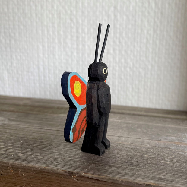 Whittled and painted wooden sculpture of a black moth, standing on 2 legs with its arms at its side and tall antennae. It has colorful red and yellow wings with a blue outline.