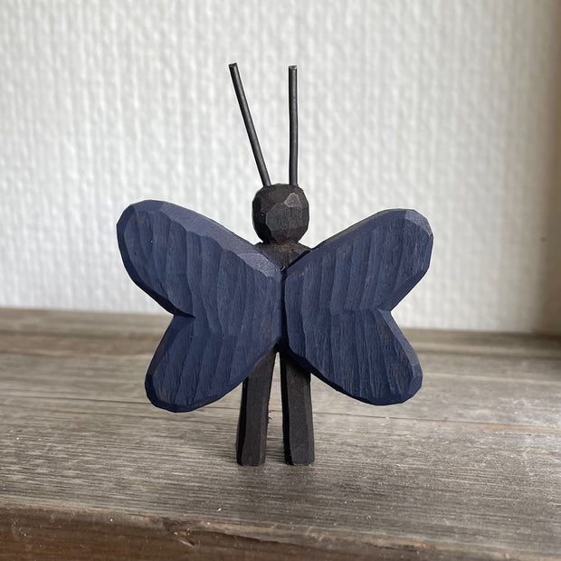 Whittled and painted wooden sculpture of a black moth, standing on 2 legs with its arms at its side and tall antennae. It has colorful red and yellow wings with a blue outline.