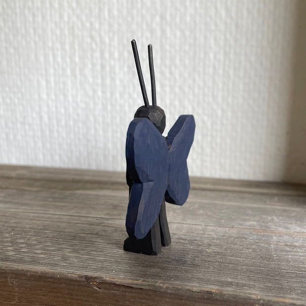 Whittled and painted wooden sculpture of a black moth, standing on 2 legs with its arms at its side and tall antennae. It has colorful red and yellow wings with a blue outline.