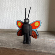 Whittled and painted wooden sculpture of a black moth, standing on 2 legs with its arms at its side and tall antennae. It has colorful red and yellow wings with a blue outline.