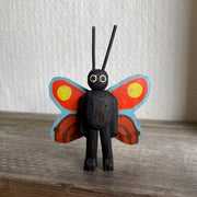 Whittled and painted wooden sculpture of a black moth, standing on 2 legs with its arms at its side and tall antennae. It has colorful red and yellow wings with a blue outline.