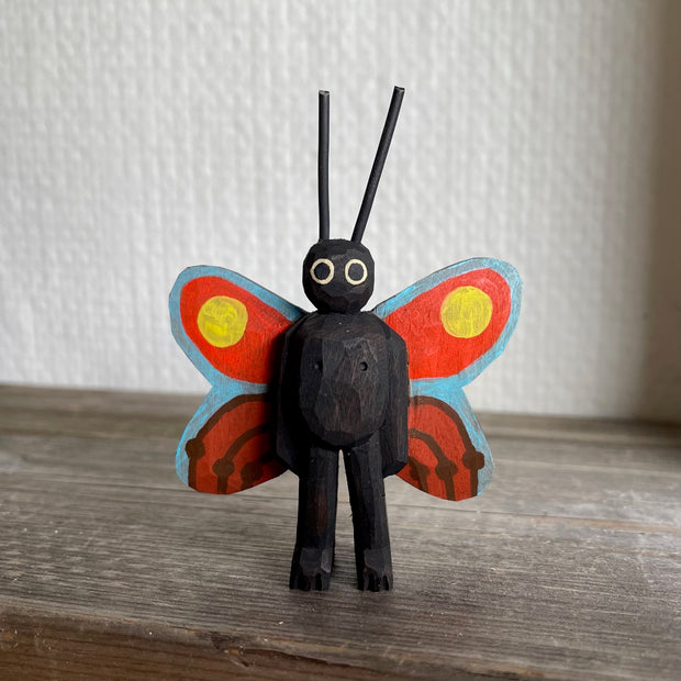 Whittled and painted wooden sculpture of a black moth, standing on 2 legs with its arms at its side and tall antennae. It has colorful red and yellow wings with a blue outline.