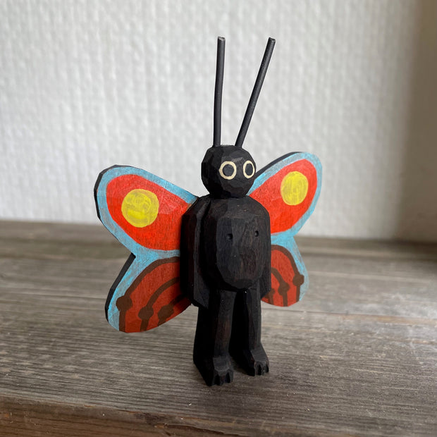 Whittled and painted wooden sculpture of a black moth, standing on 2 legs with its arms at its side and tall antennae. It has colorful red and yellow wings with a blue outline.