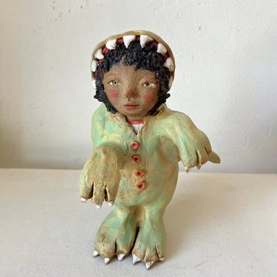 Ceramic sculpture of a tan girl with curly hair dressed up in a fully body dinosaur costume, with an open dinosaur mouth for the hood. She has a curious expression.