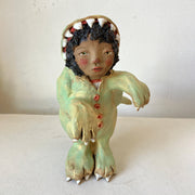 Ceramic sculpture of a tan girl with curly hair dressed up in a fully body dinosaur costume, with an open dinosaur mouth for the hood. She has a curious expression.