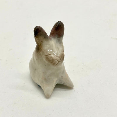 Ceramic sculpture of a rabbit, sitting with its front legs spread out in front and pointed. It has dark brown ears on a white body and a closed eye expression with an "x" shape making up its nose and mouth.