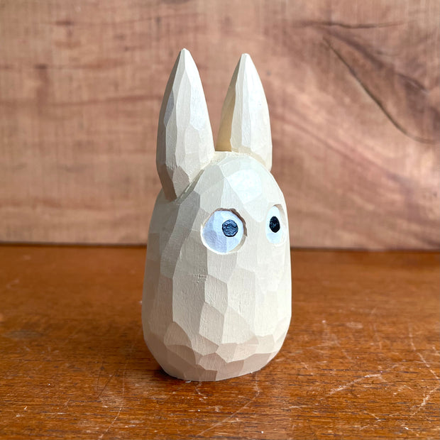 Whittled wooden sculpture of a small, off white ghost creature with large cartoon eyes, tall pointed ears and no limbs or other facial features. It looks straight ahead.