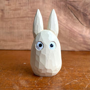 Whittled wooden sculpture of a small, off white ghost creature with large cartoon eyes, tall pointed ears and no limbs or other facial features. It looks straight ahead.