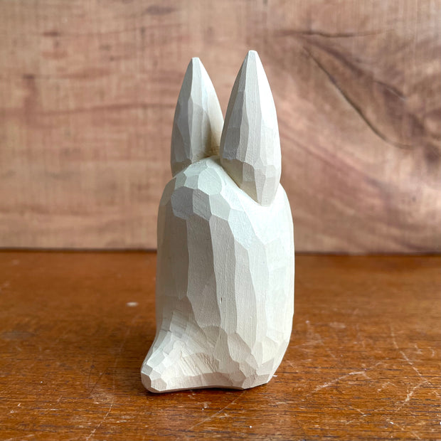 Whittled wooden sculpture of a small, off white ghost creature with large cartoon eyes, tall pointed ears and no limbs or other facial features. It looks straight ahead.