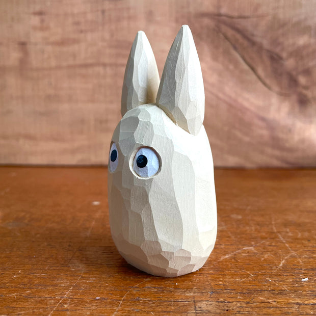 Whittled wooden sculpture of a small, off white ghost creature with large cartoon eyes, tall pointed ears and no limbs or other facial features. It looks straight ahead.