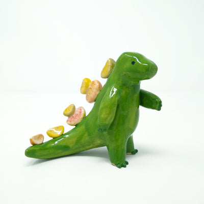 Ceramic sculpture of a green Godzilla like monster with smooth limbs and a simplistic face. Lining its back like spikes are slices of oranges and lemons.