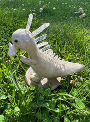 Ceramic sculpture of a cream colored dinosaur with large quartz on its back like spikes. It dangles a small cute character from its mouth by their foot.