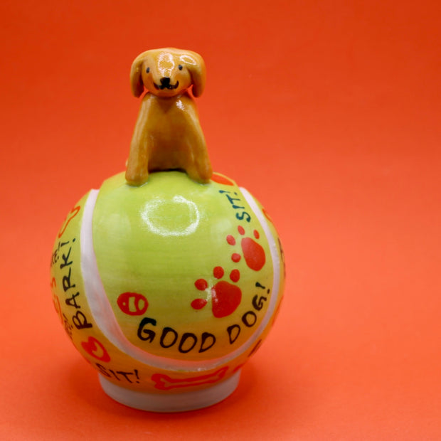 Ceramic sculpture of a small brown dog smiling and sitting atop a large tennis ball, with affirmations written on it with cute dog themed doodles.