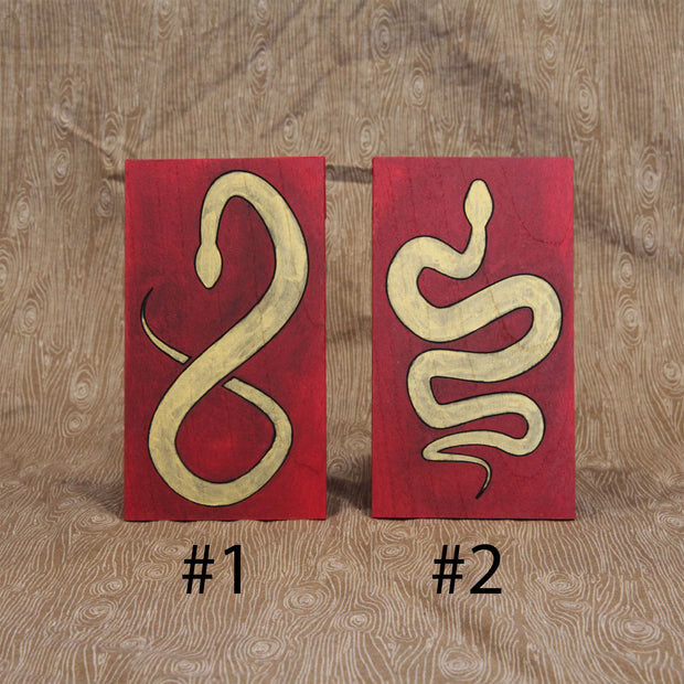 2 red envelopes made out of wood, with painted illustrations on each of gold snakes with black outlines. One snake is curved into itself, akin to the shape of an 8. The other is curved to make a multi-part "S" shape.