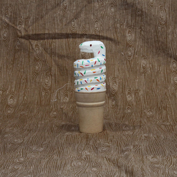 Carved wooden sculpture of a sugar style ice cream cone, holding a curved scoop of white soft serve shaped like a coiled snake. It has rainbow colored sprinkles atop its body.