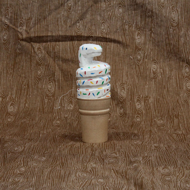 Carved wooden sculpture of a sugar style ice cream cone, holding a curved scoop of white soft serve shaped like a coiled snake. It has rainbow colored sprinkles atop its body.