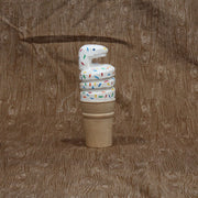 Carved wooden sculpture of a sugar style ice cream cone, holding a curved scoop of white soft serve shaped like a coiled snake. It has rainbow colored sprinkles atop its body.