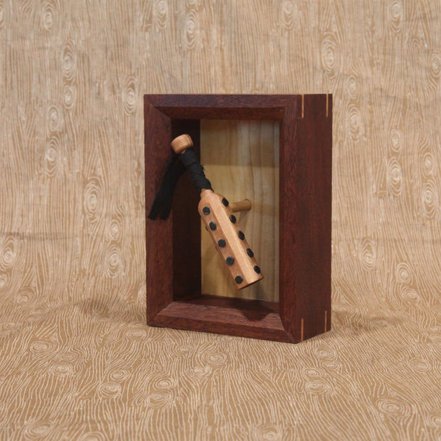 Wooden sculpture of a wooden bat, studded with metal circles and a cloth wrapped handle. It is contained in a shadowbox style wood frame.