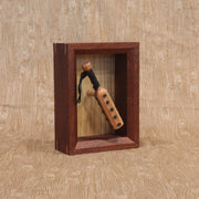 Wooden sculpture of a wooden bat, studded with metal circles and a cloth wrapped handle. It is contained in a shadowbox style wood frame.