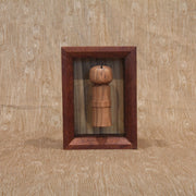 Sculpture of a wooden kokeshi, with a single spike coming out the top. It is encased in a shadowbox style wooden frame.