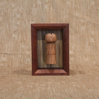 Sculpture of a wooden kokeshi, with a single spike coming out the top. It is encased in a shadowbox style wooden frame.