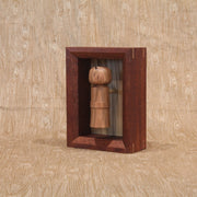 Sculpture of a wooden kokeshi, with a single spike coming out the top. It is encased in a shadowbox style wooden frame.