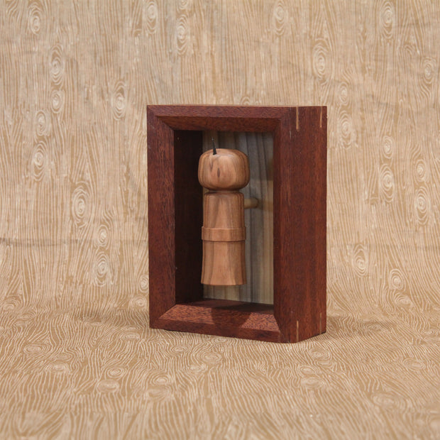 Sculpture of a wooden kokeshi, with a single spike coming out the top. It is encased in a shadowbox style wooden frame.