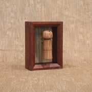 Sculpture of a wooden kokeshi, with a single spike coming out the top. It is encased in a shadowbox style wooden frame.