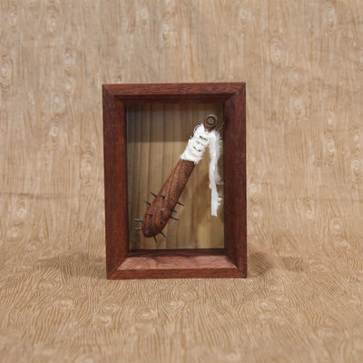 Sculpture of a small wooden bat with spikes coming out the top. A white strip of fabric is wrapped around the handle. Piece is within a shadowbox style wooden frame.