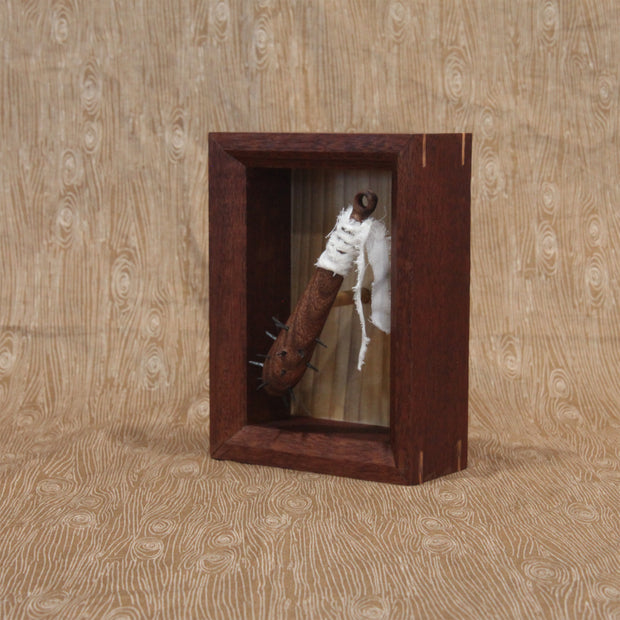 Sculpture of a small wooden bat with spikes coming out the top. A white strip of fabric is wrapped around the handle. Piece is within a shadowbox style wooden frame.