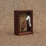 Sculpture of a small wooden bat with spikes coming out the top. A white strip of fabric is wrapped around the handle. Piece is within a shadowbox style wooden frame.